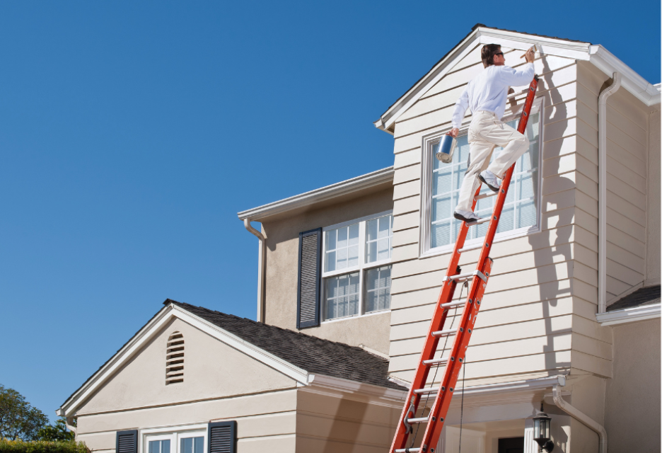 A contractor on an extension ladder paints the exterior siding near an upper-story window of a house as part of fall home maintenance.